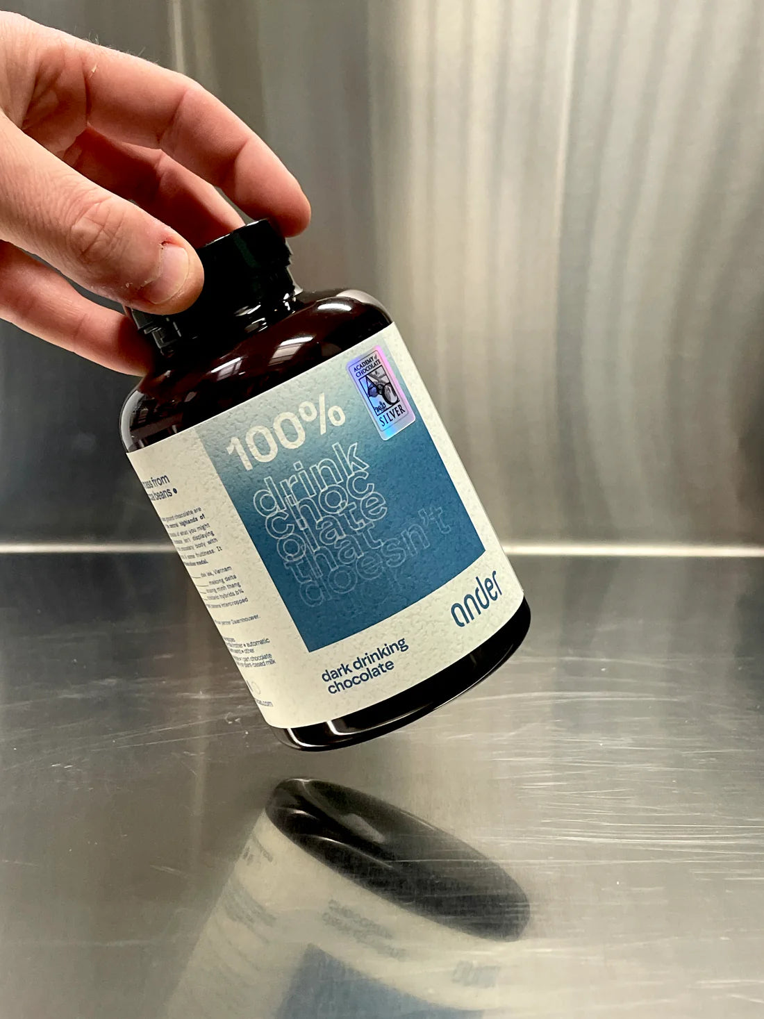 Angled view of a hand holding a bottle of Ander 100% Dark Drinking Chocolate against a metallic background.