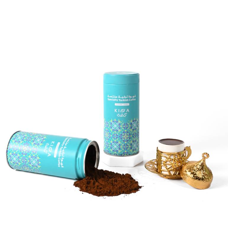 Lifestyle shot of Kiffa Specialty Turkish Coffee turquoise tins, one upright and one tipped over with ground coffee spilled on a white surface next to a gold cup.